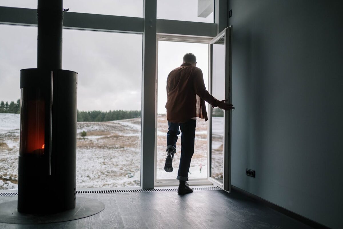 Man exiting modern home onto snowy landscape through glass door with a fireplace inside.